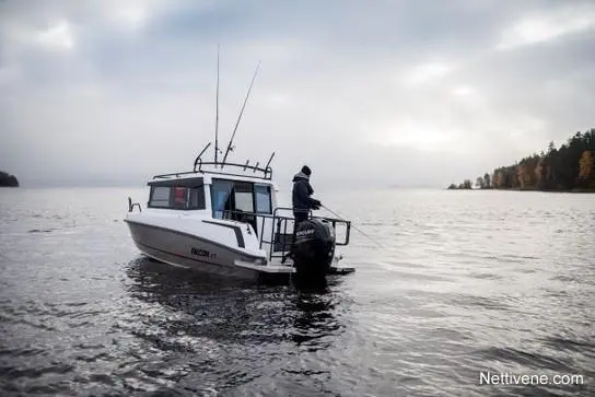 A man stands on a fishing boat in calm waters, with a forested shoreline under a cloudy sky.